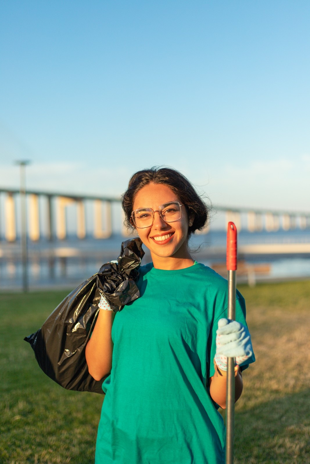 A smiling young woman wearing gloves and a teal shirt holds a trash bag and cleaning tool in a park near a bridge, proudly participating in a volunteer travel for beginners cleanup activity.