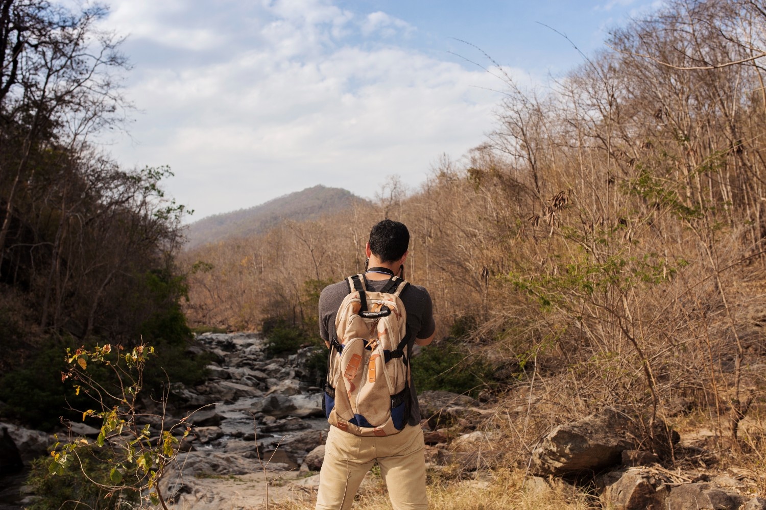 A traveler with a backpack stands near a rocky stream surrounded by dry forest and hills, capturing the natural landscape with his camera a peaceful adventure moment perfect for a first time volunteer exploring nature and sustainable travel opportunities.