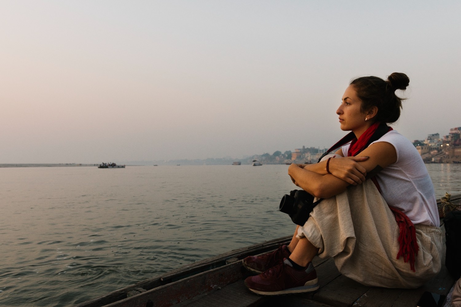 A woman sitting peacefully on a wooden boat at sunrise on the Ganges River, holding a camera and gazing at the distant shore a serene and reflective moment symbolizing the beauty of travel for free in India.