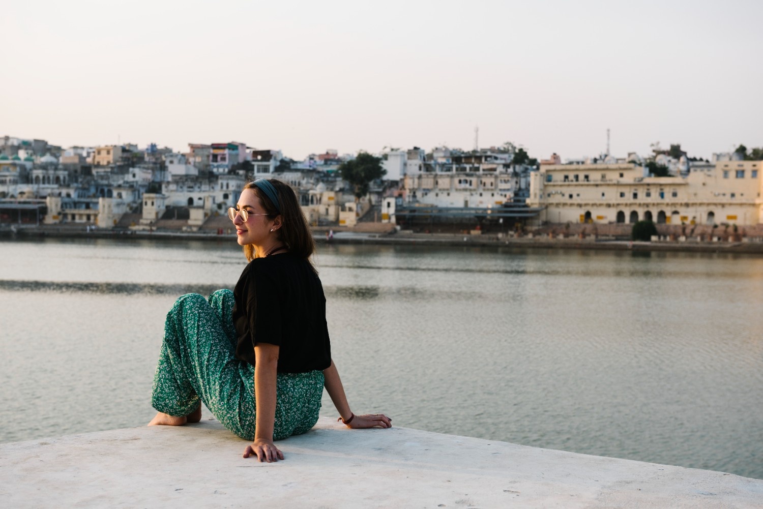 A young woman sitting peacefully by a lake, enjoying the scenic view of traditional Indian architecture and calm waters at sunset a serene moment that reflects the cultural beauty and personal growth found during a volunteer travel experience in India.